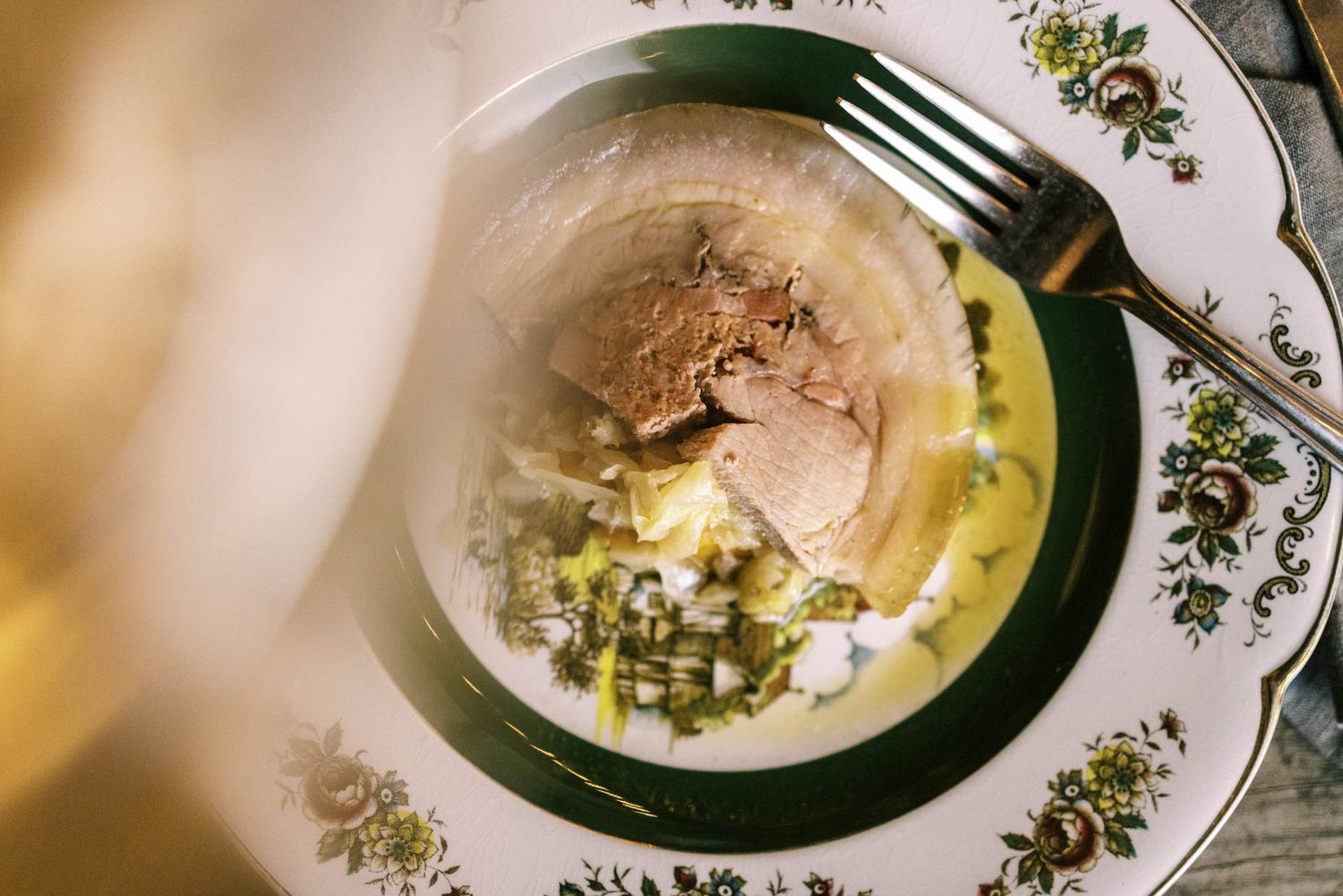 Elegant plate with sliced meat, creamy sauce, and herb garnish on ornate green and white china with a silver fork.