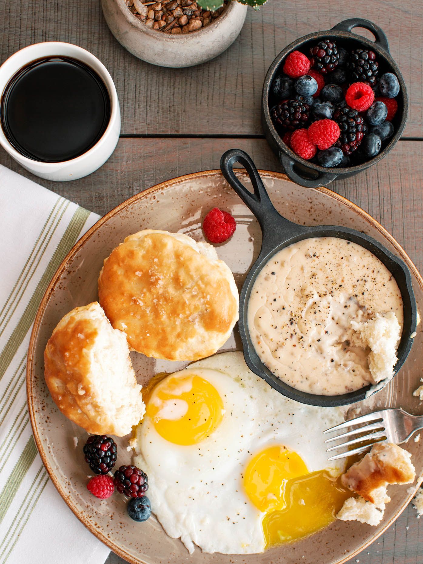 Classic Southern breakfast with fried eggs, biscuits, sausage gravy, fresh berries, and black coffee on a rustic plate.