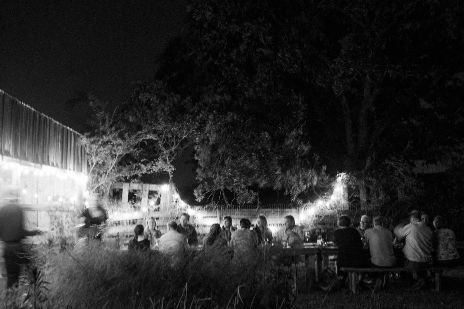 Black and white photo of people dining outdoors at night under string lights and trees at a garden gathering.