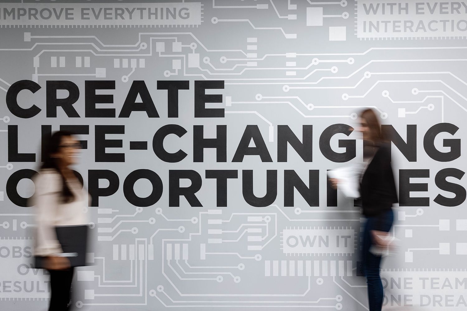 Two professionals walk past a large wall mural reading Create Life-Changing Opportunities with circuit board design.