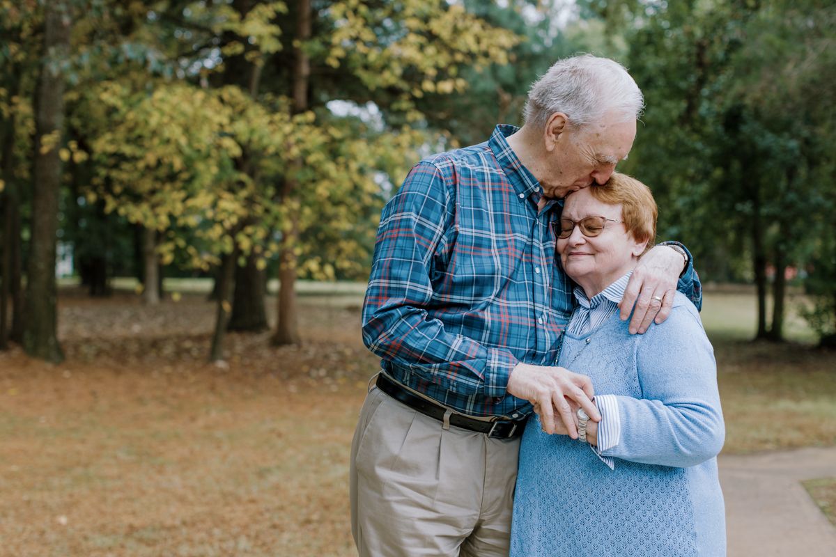 Elderly couple embracing outdoors in a park, man kissing woman on head, both smiling warmly among autumn trees.