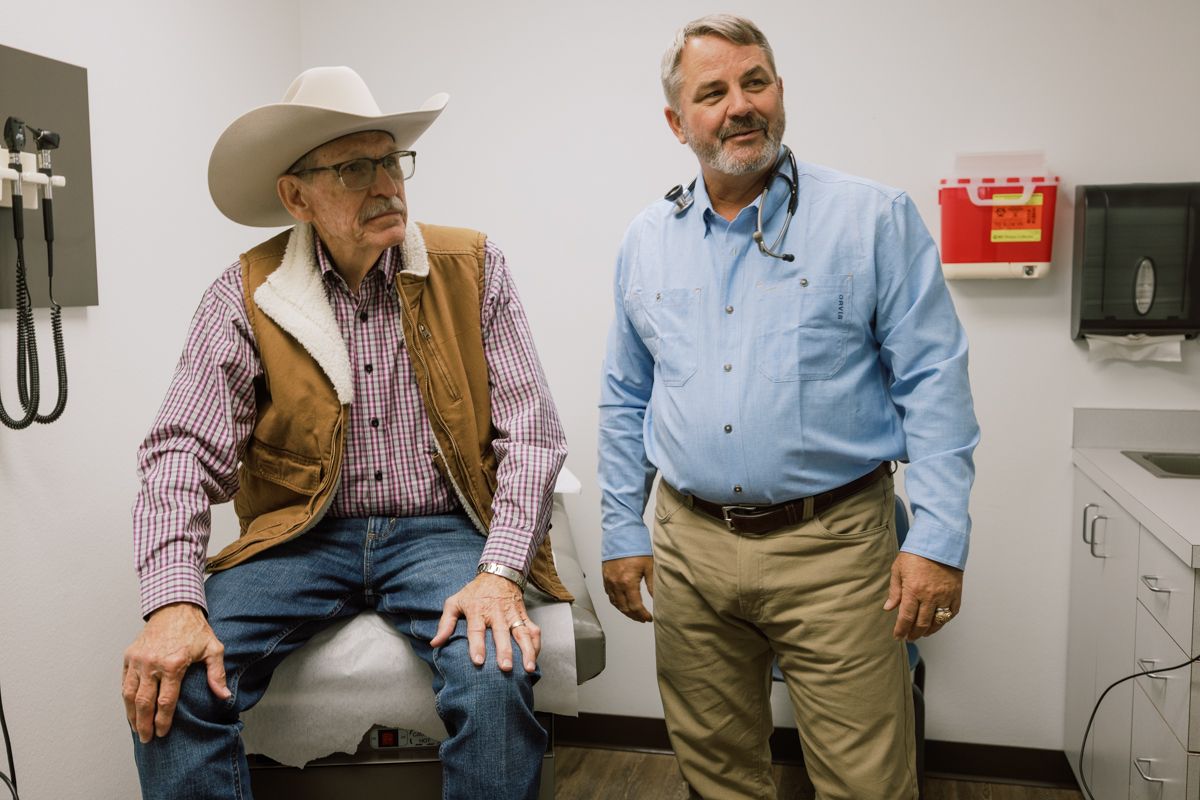 A doctor in a blue shirt speaks with an elderly patient wearing a cowboy hat in an exam room.