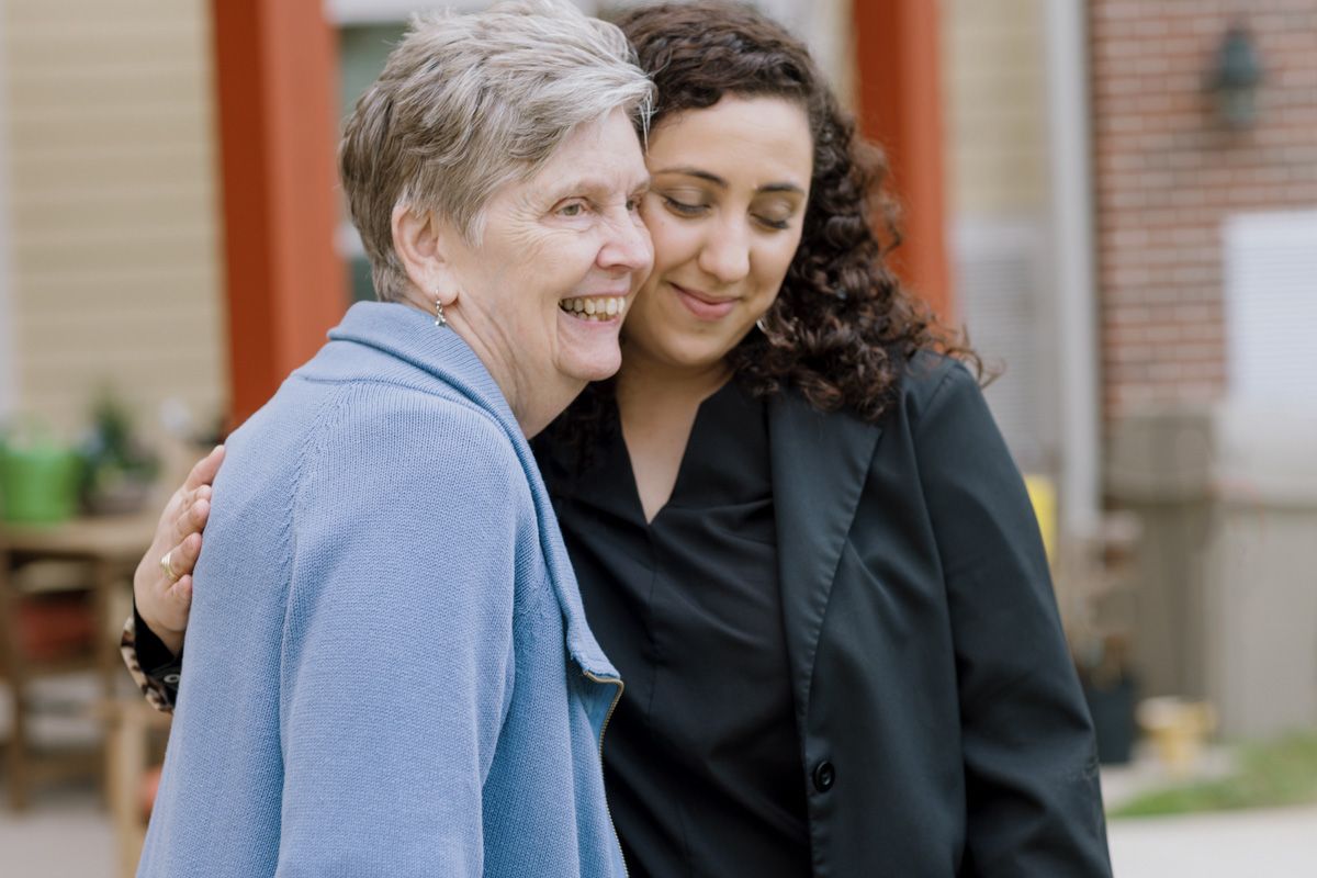 A young woman in black gently embraces a smiling elderly woman in a blue cardigan outdoors.