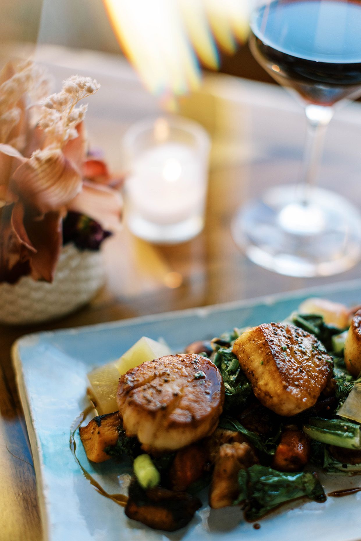 Seared scallops on blue plate with vegetables, candlelight, and red wine glass in elegant restaurant setting.