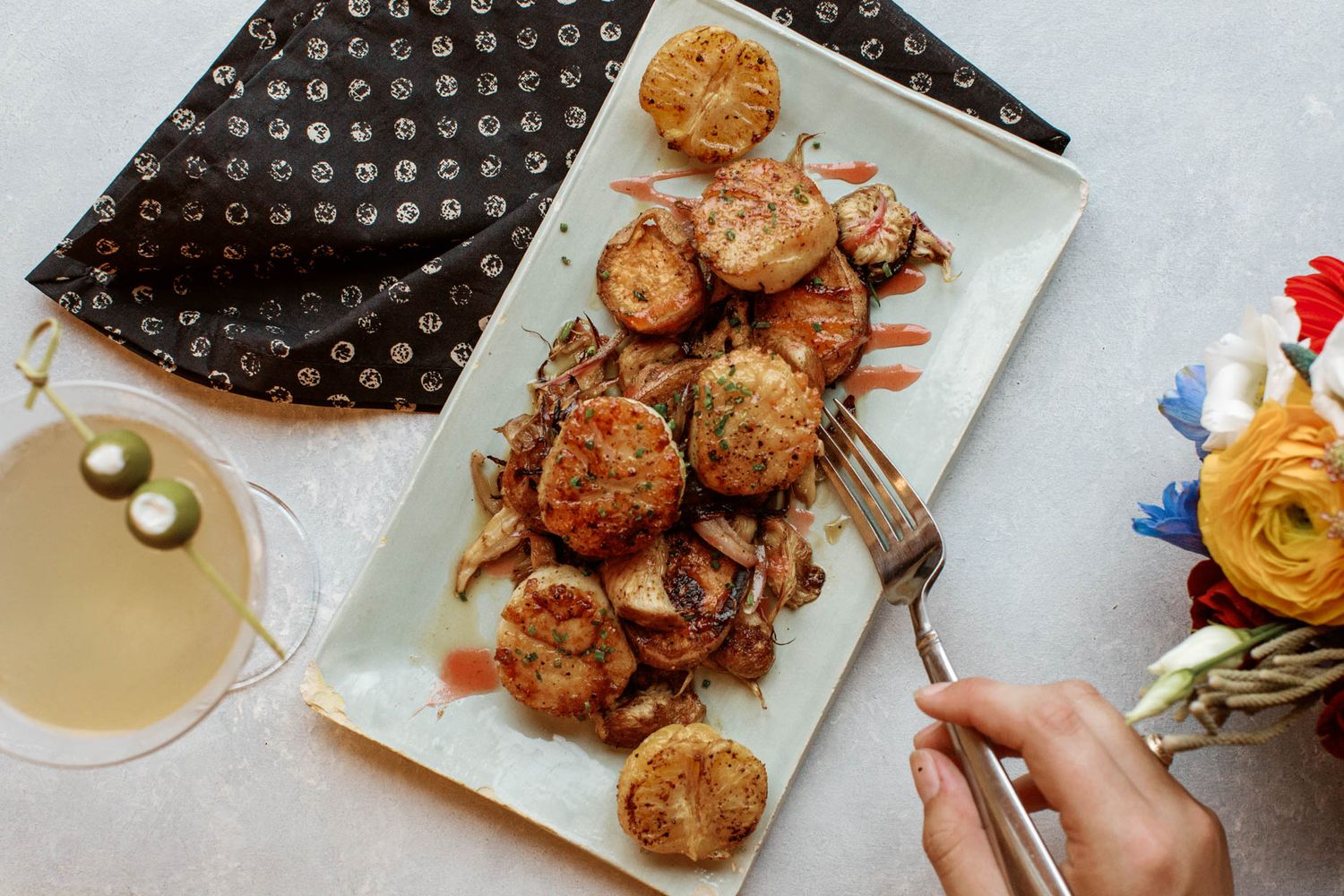Overhead view of seared scallops on a white rectangular plate with a cocktail and flowers nearby.