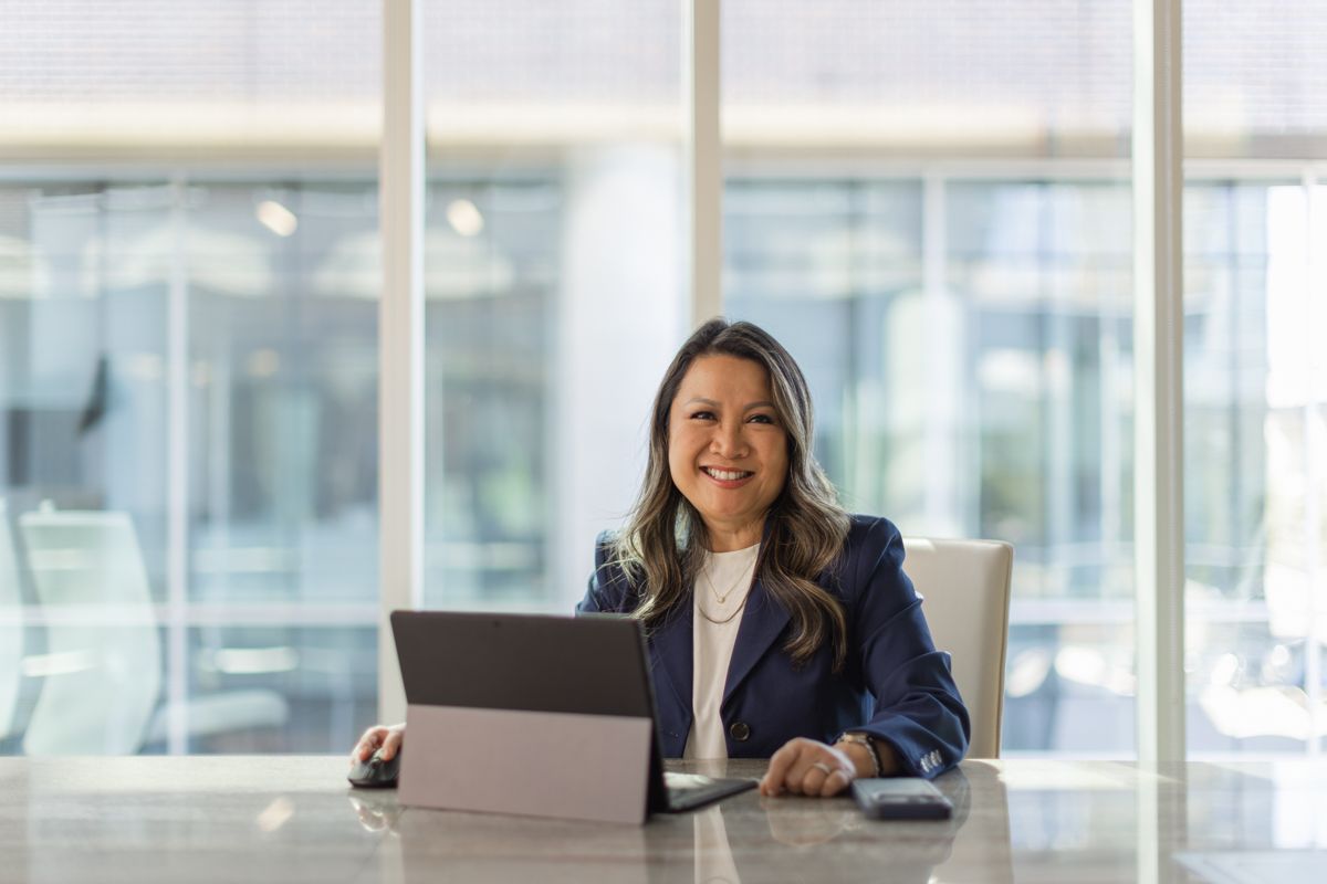 Confident businesswoman in navy blazer smiling while working on laptop at modern office desk.