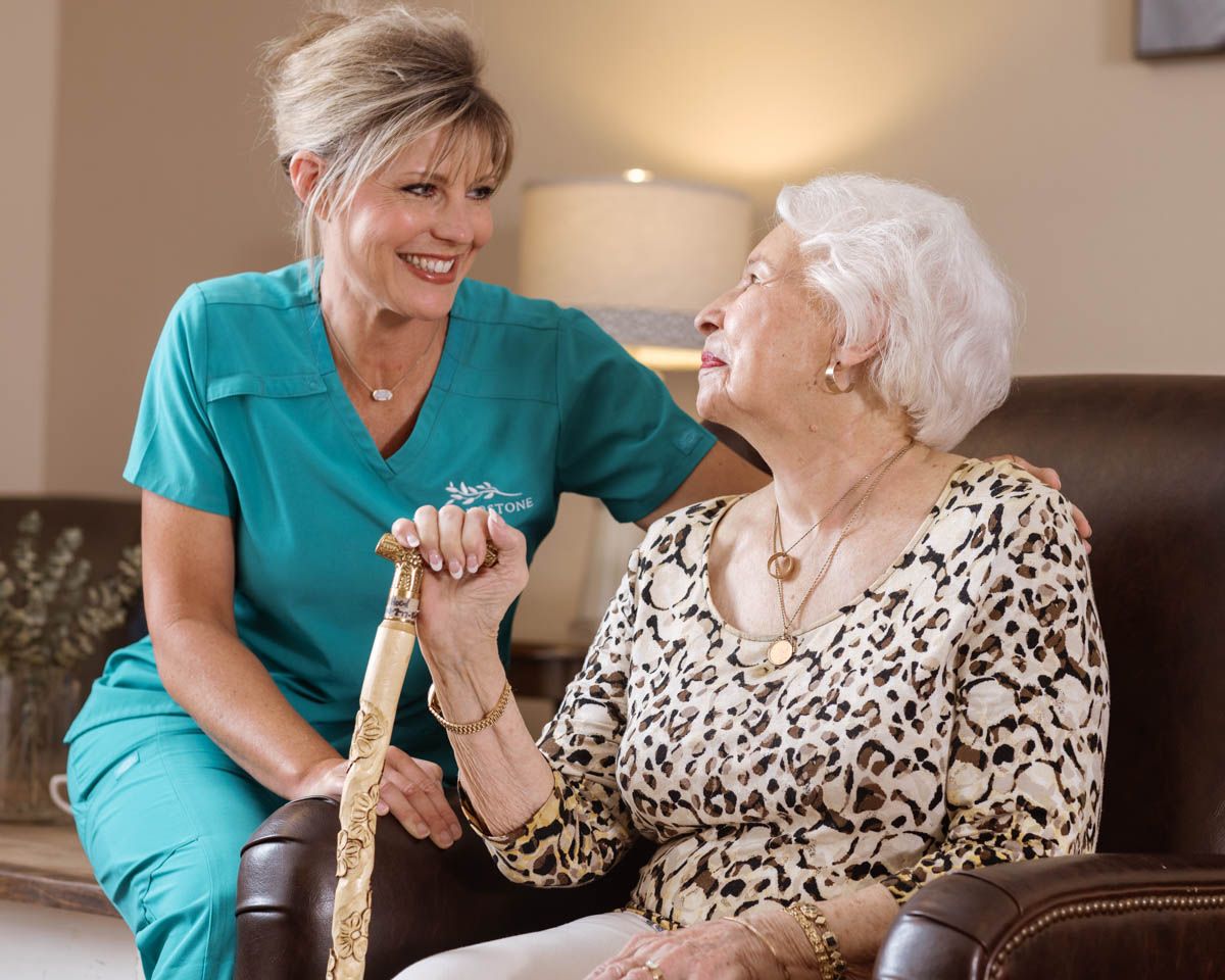 Caring nurse in teal scrubs smiles warmly with elderly resident holding a decorative cane indoors.