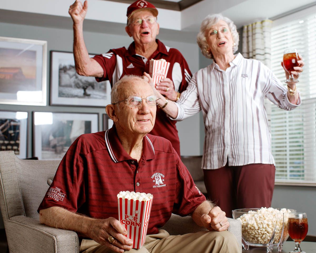 Three elderly fans in maroon shirts cheer excitedly with popcorn and drinks while watching a sports game together indoors.