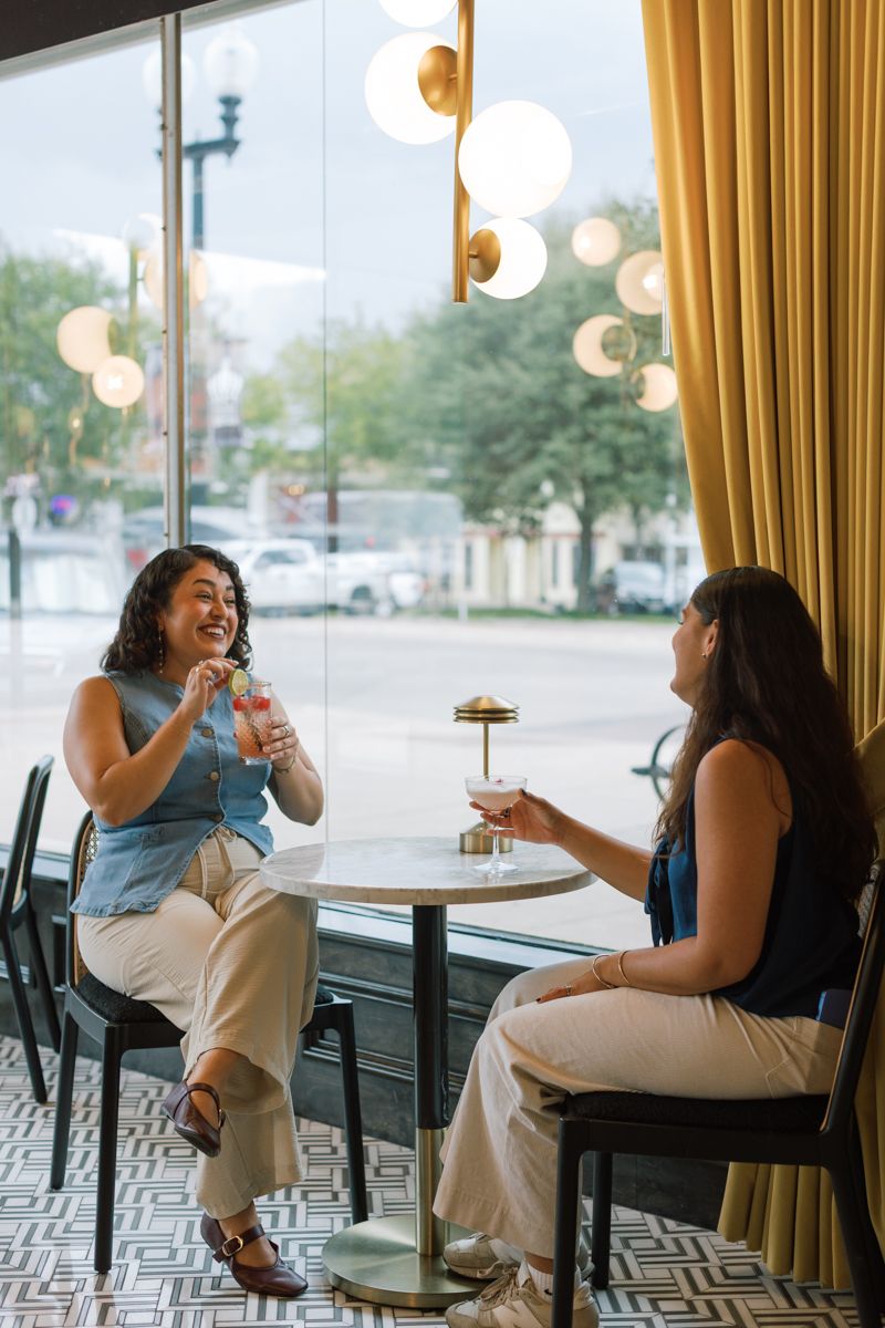 Two women laughing and enjoying cocktails at a stylish bar table by a large window with golden curtains.