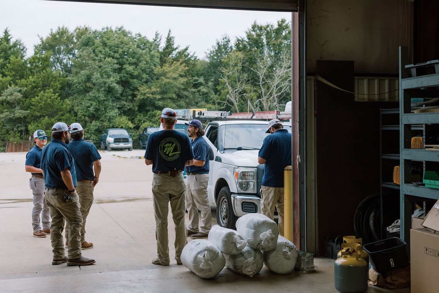 A crew of workers in blue uniforms gather near a white truck inside an open garage bay with supplies nearby.