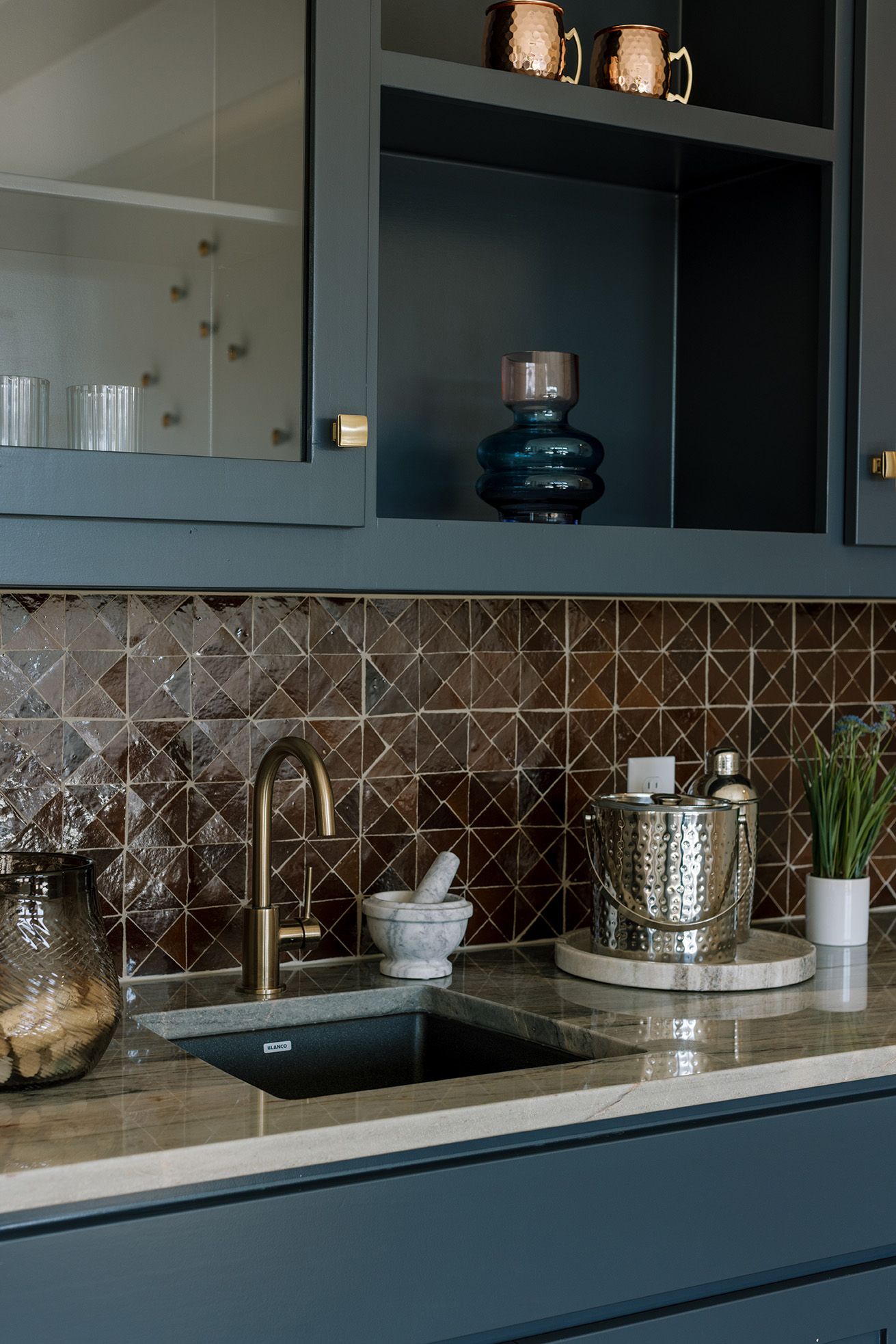 Dark teal wet bar with geometric brown tile backsplash, brass faucet, marble countertop, and glass cabinet doors.