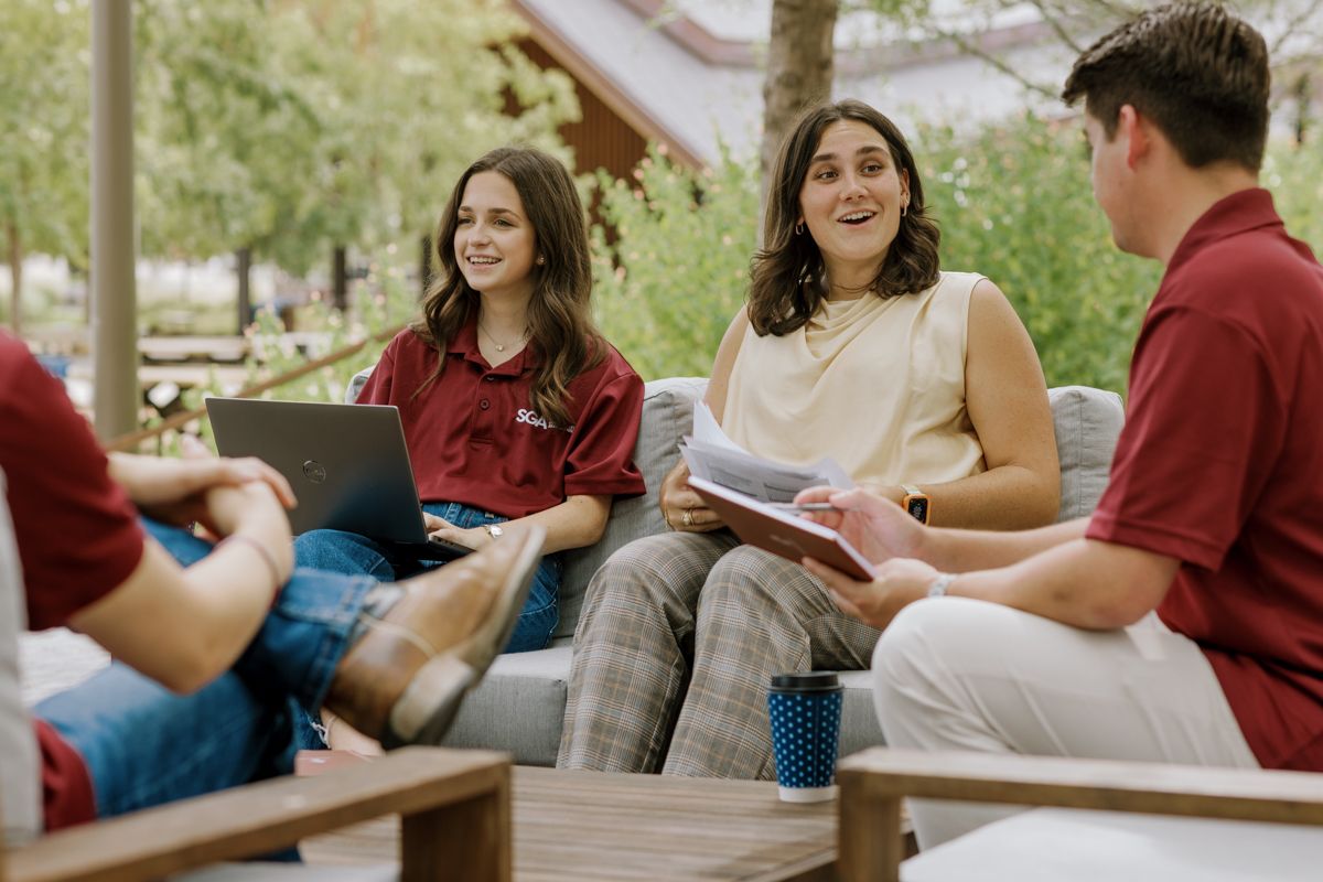 Four college students laughing and conversing while seated on outdoor patio furniture with laptops and books.