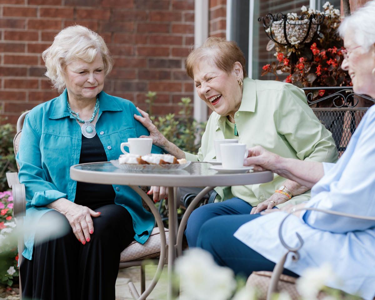 Three senior women laughing and enjoying coffee together at an outdoor patio table.
