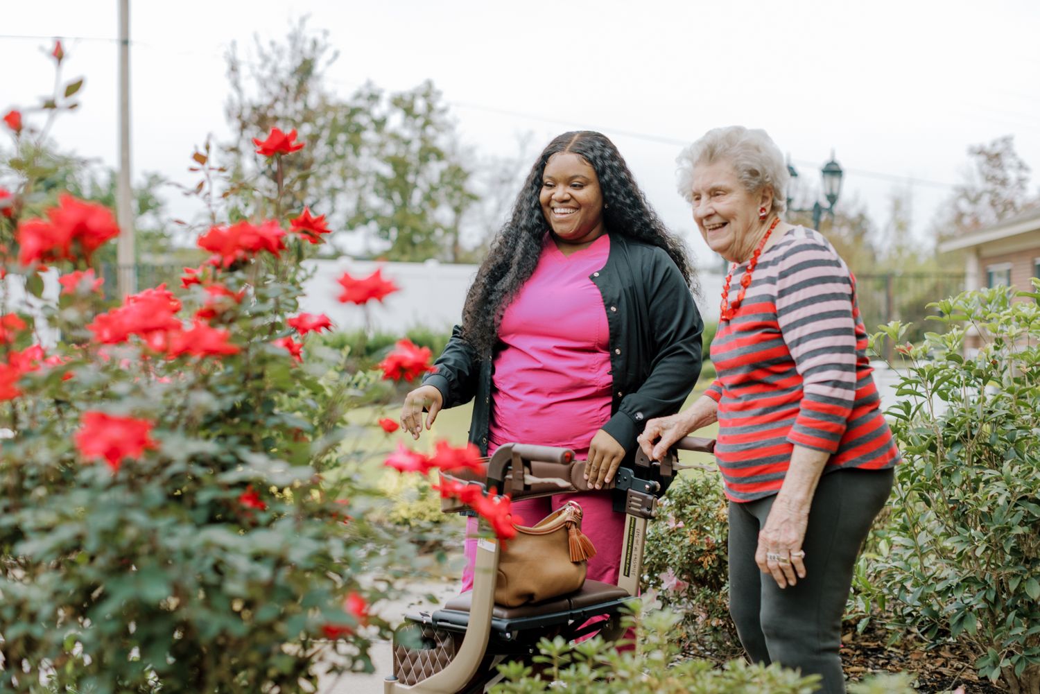 A young Black caregiver and elderly white woman smile together among red roses outdoors, with a walker nearby.