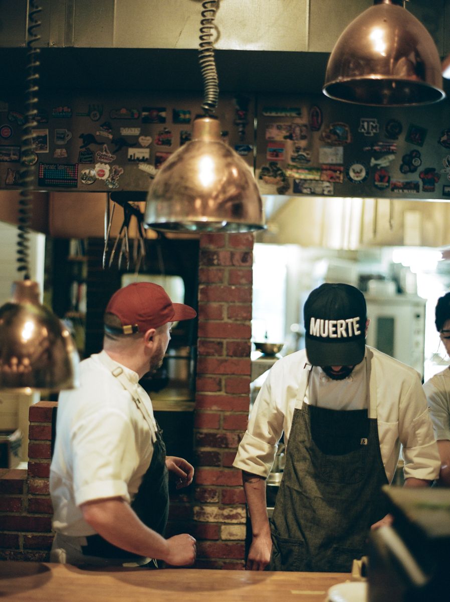Two chefs working in a rustic kitchen under copper pendant lights, one wearing a Muerte cap.