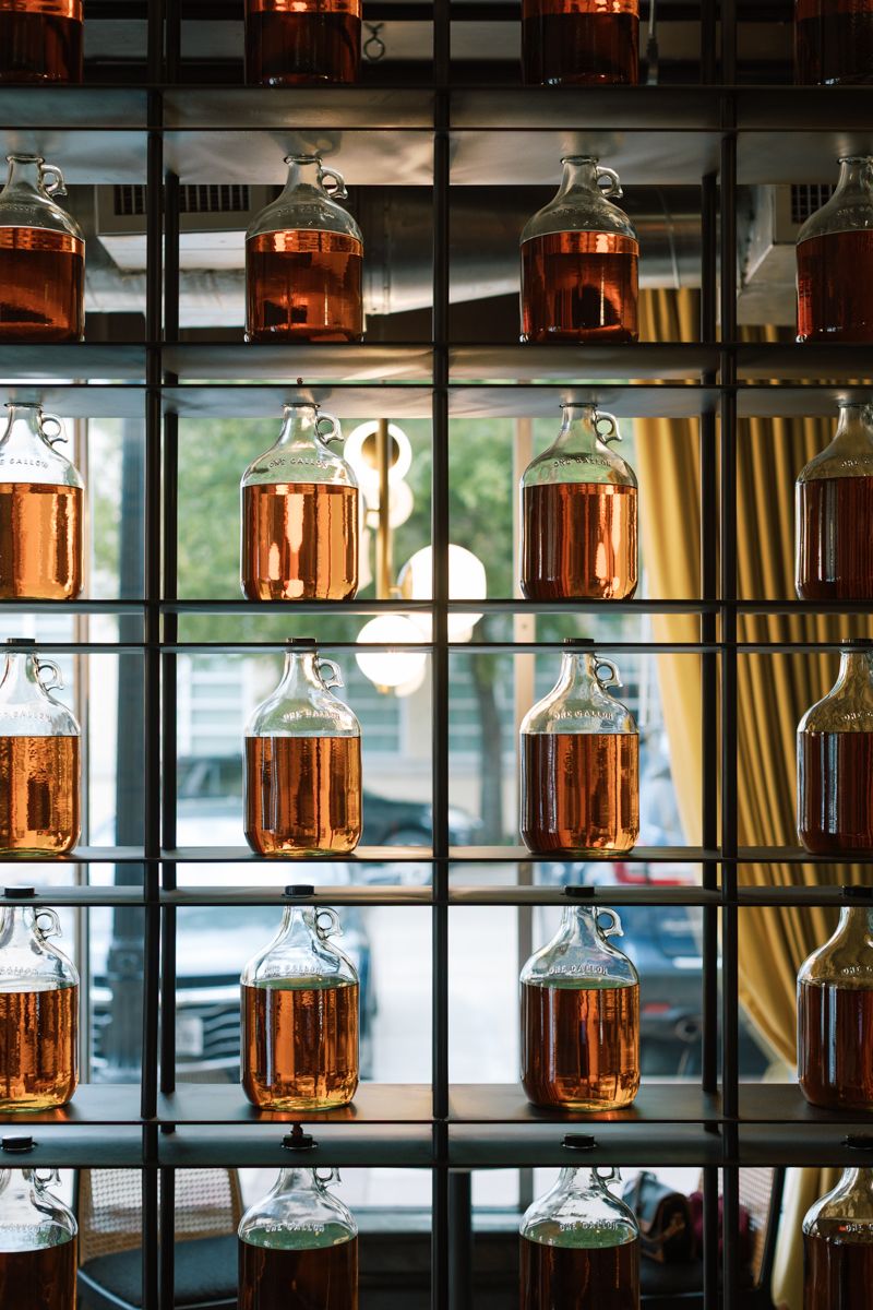 Rows of large glass jugs filled with golden amber liquid displayed on metal shelving unit in front of a window.