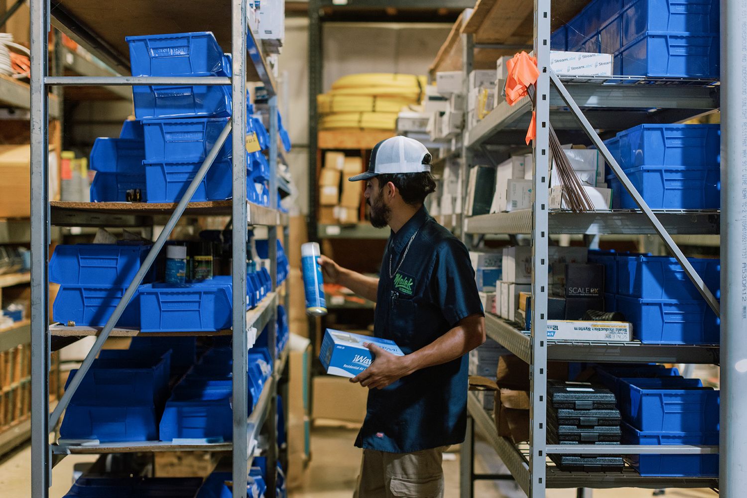 Worker in white cap organizing blue bins on metal shelving in a warehouse storage facility.