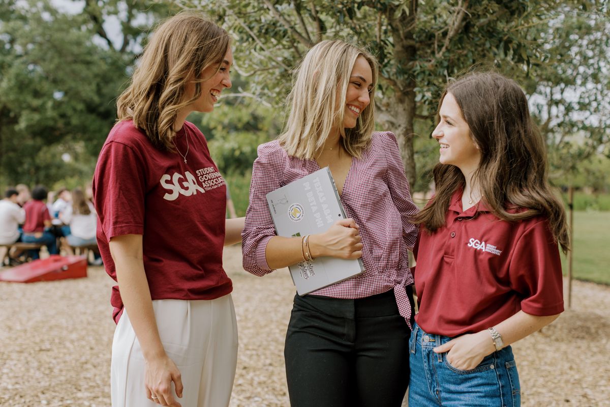 Three smiling women in SGA and university polo shirts chatting outdoors at a campus gathering event.