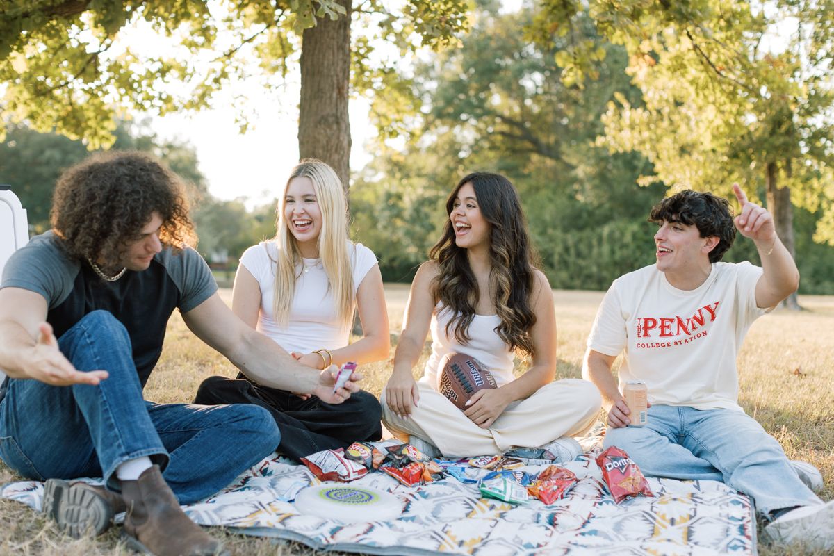 Four young friends laughing and enjoying a sunny outdoor picnic on a blanket under a tree in a park.