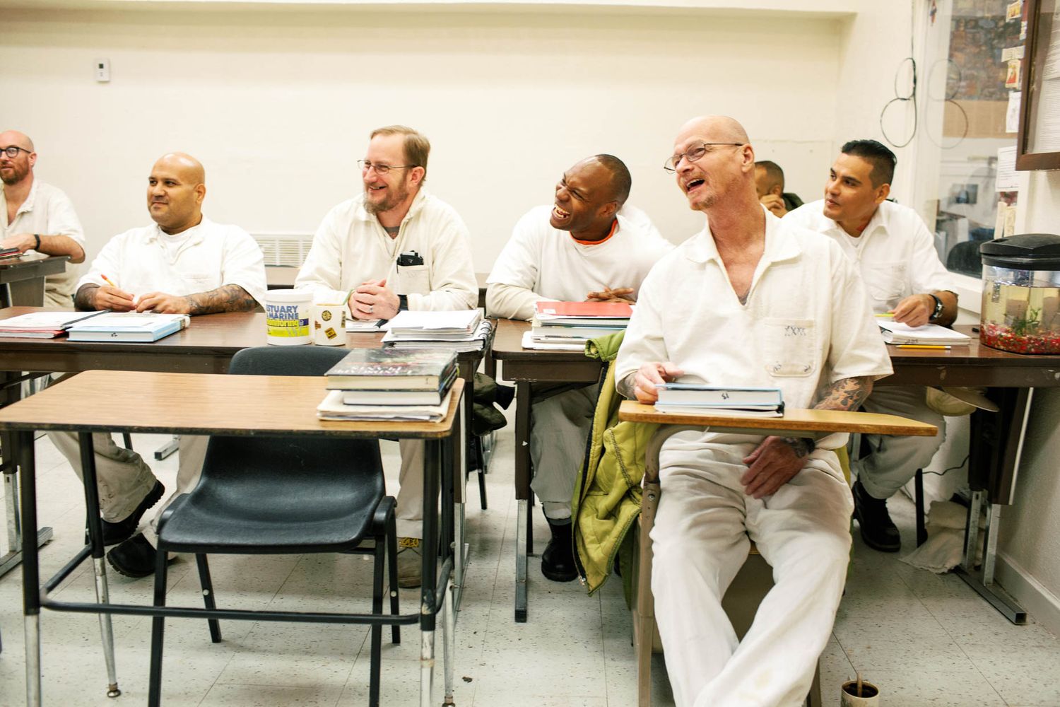 Group of men in white clothing laughing and smiling while sitting at desks in a classroom setting.