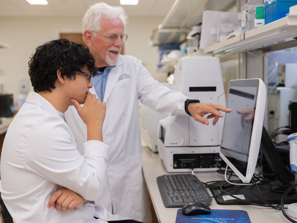 Two scientists in white lab coats collaborate while reviewing data on a computer monitor in a lab.