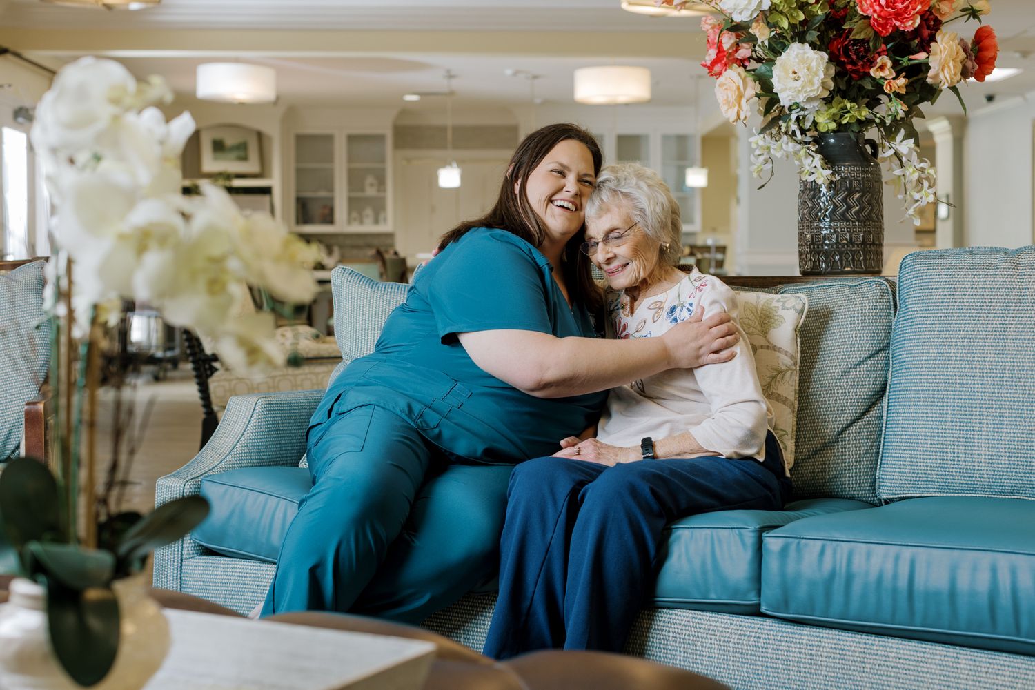 A caregiver in teal scrubs warmly hugs an elderly woman on a light blue sofa in a cozy living room.