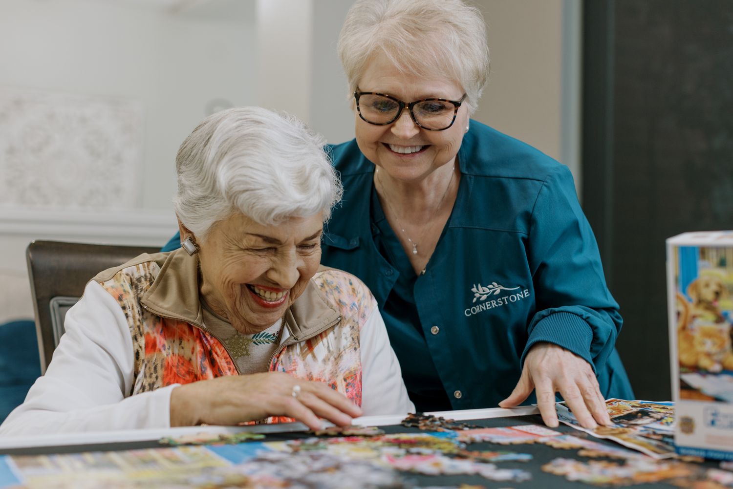 Two elderly women smiling and working together on a colorful jigsaw puzzle at a table indoors.