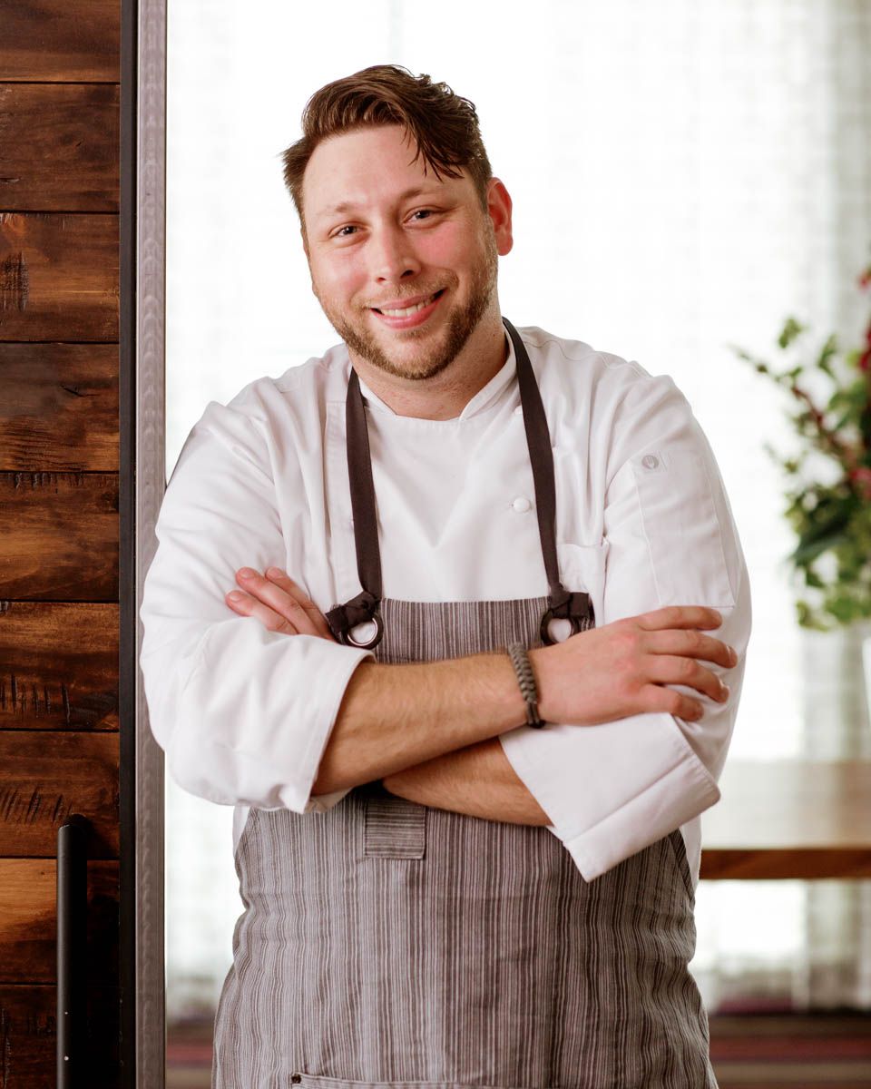 Smiling chef in white coat and striped apron stands with arms crossed in a restaurant setting.