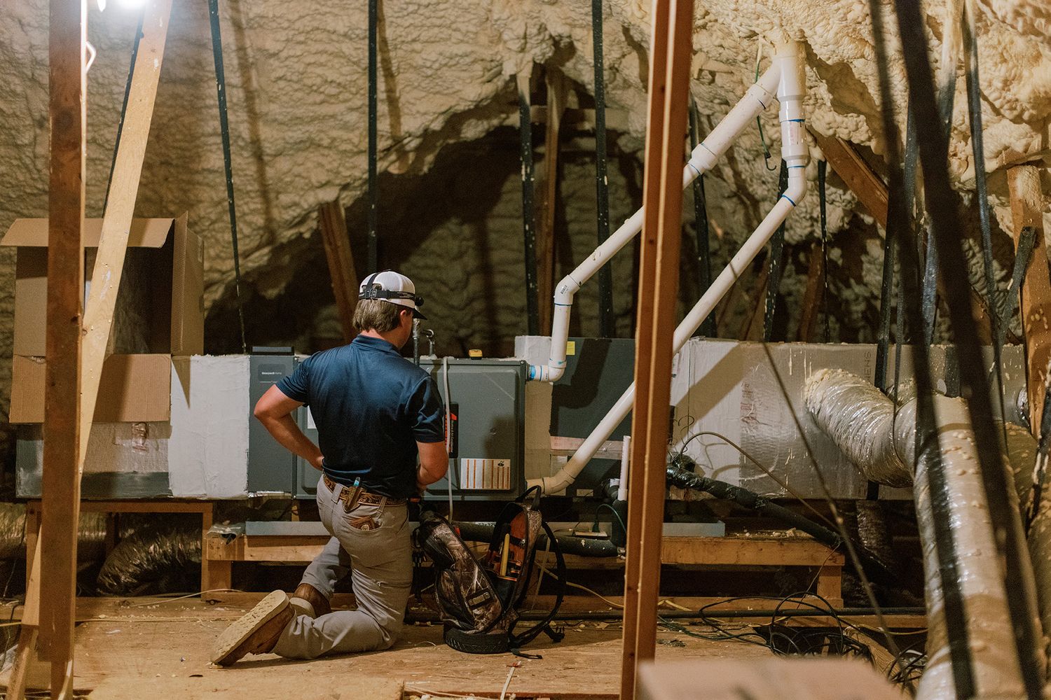 Technician kneeling in crawl space installing HVAC system surrounded by wooden framing and insulation.