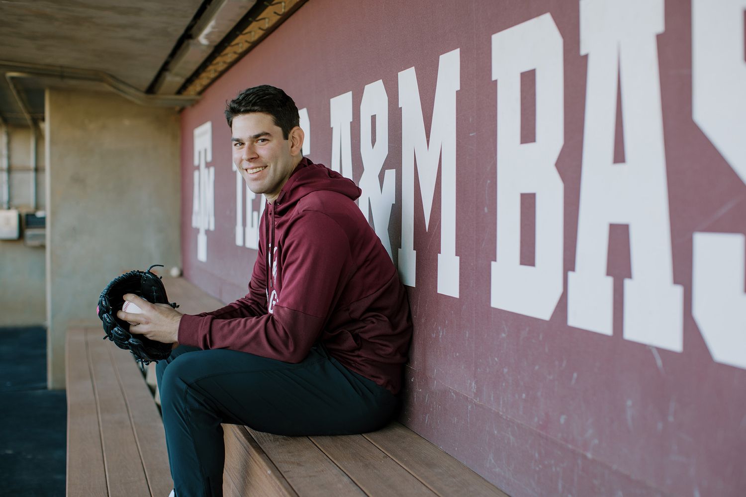 Smiling baseball player sitting in a Texas A&M dugout wearing a maroon hoodie.