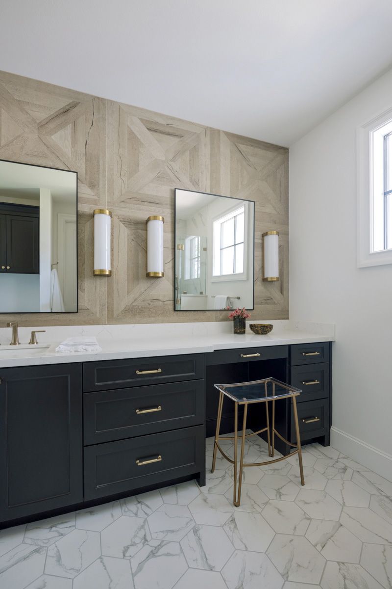 Dark navy bathroom vanity with gold hardware, geometric wood accent wall, marble floors, and dual mirrors with brass sconces.
