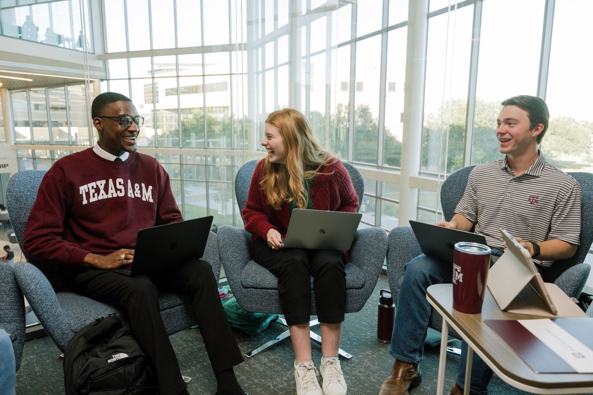 Three Texas A&M students laugh together while working on laptops in a campus common area.