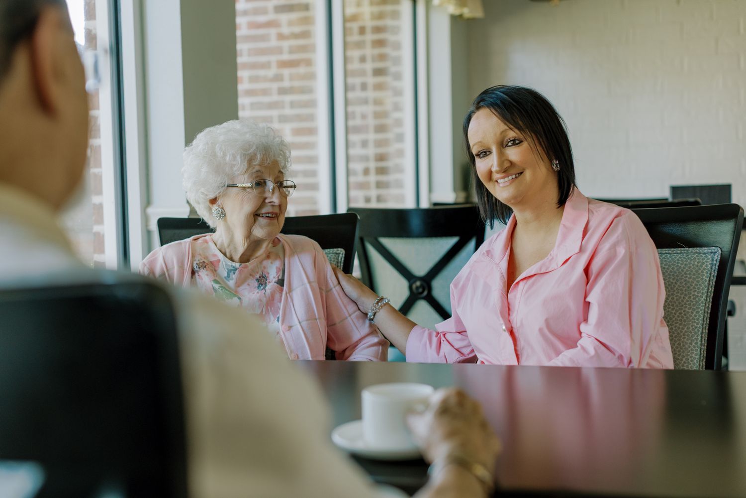 Two women in pink tops smiling and chatting at a cafe table with a white coffee cup.