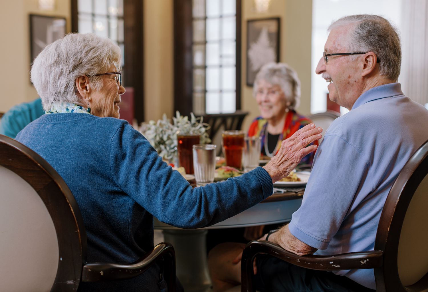 Senior couple holding hands and smiling at a restaurant table with friends in background.