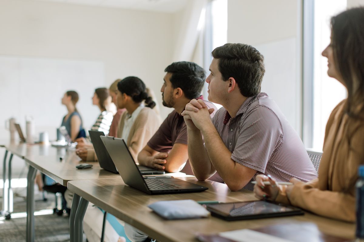Students sitting at a long desk with laptops attentively listening in a bright classroom.