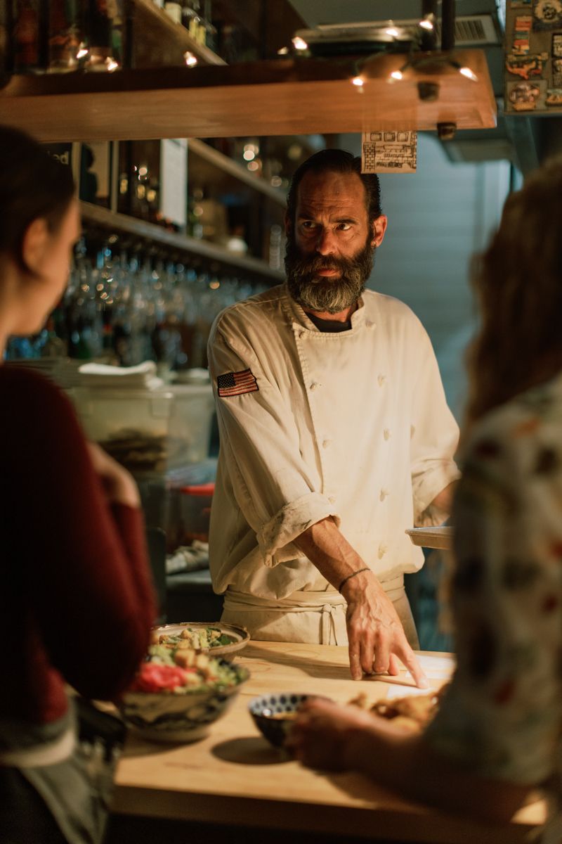 Bearded chef in white coat presenting dishes to guests at a cozy, warmly lit restaurant counter.