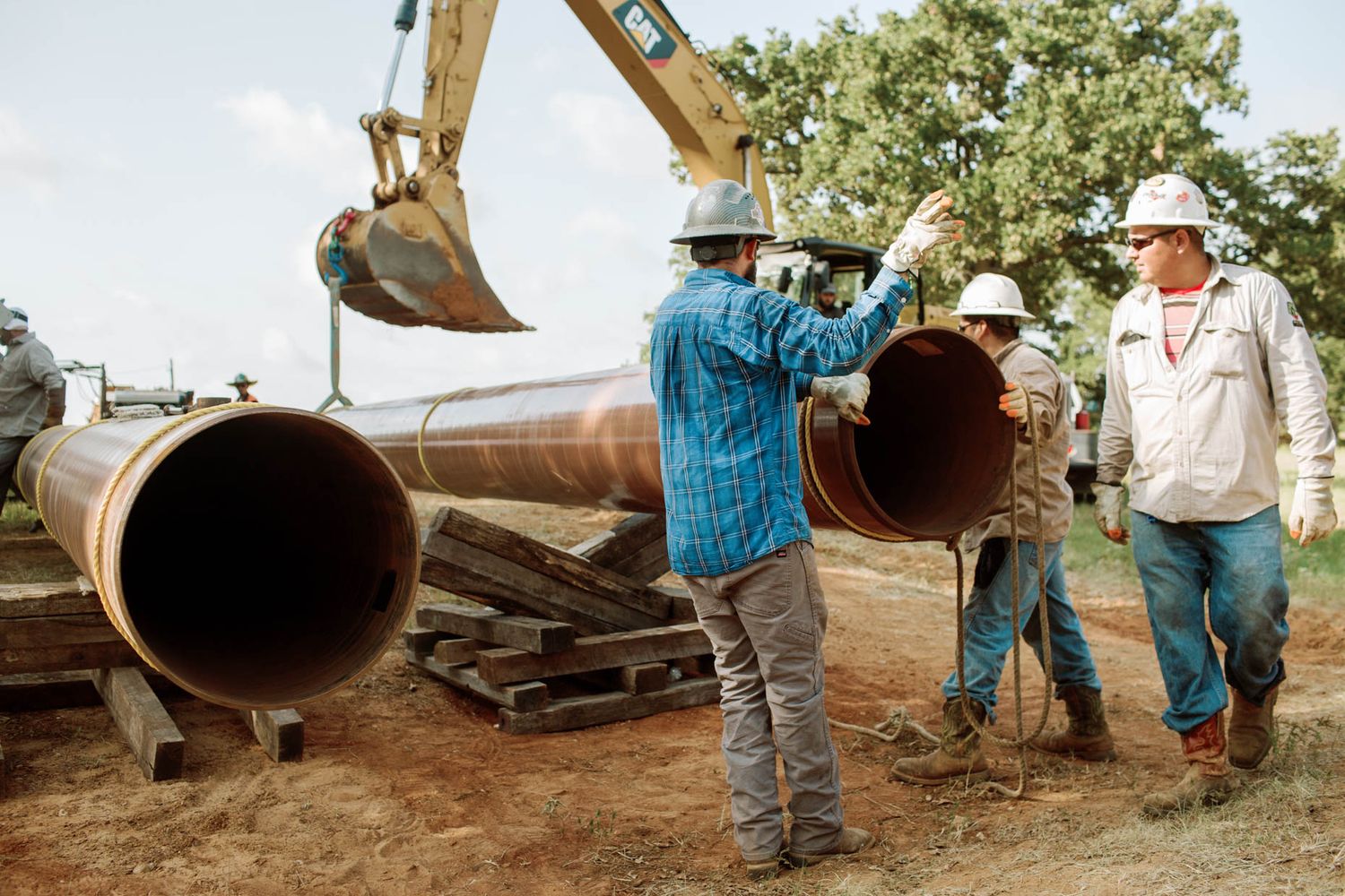 Construction workers in hard hats handling large steel pipes at an outdoor pipeline installation site with excavator.