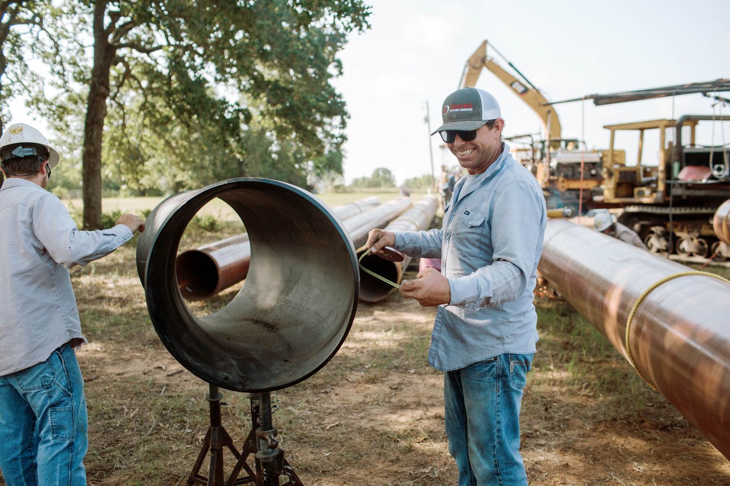 Workers handling large industrial pipes outdoors at a construction site with heavy machinery in the background.