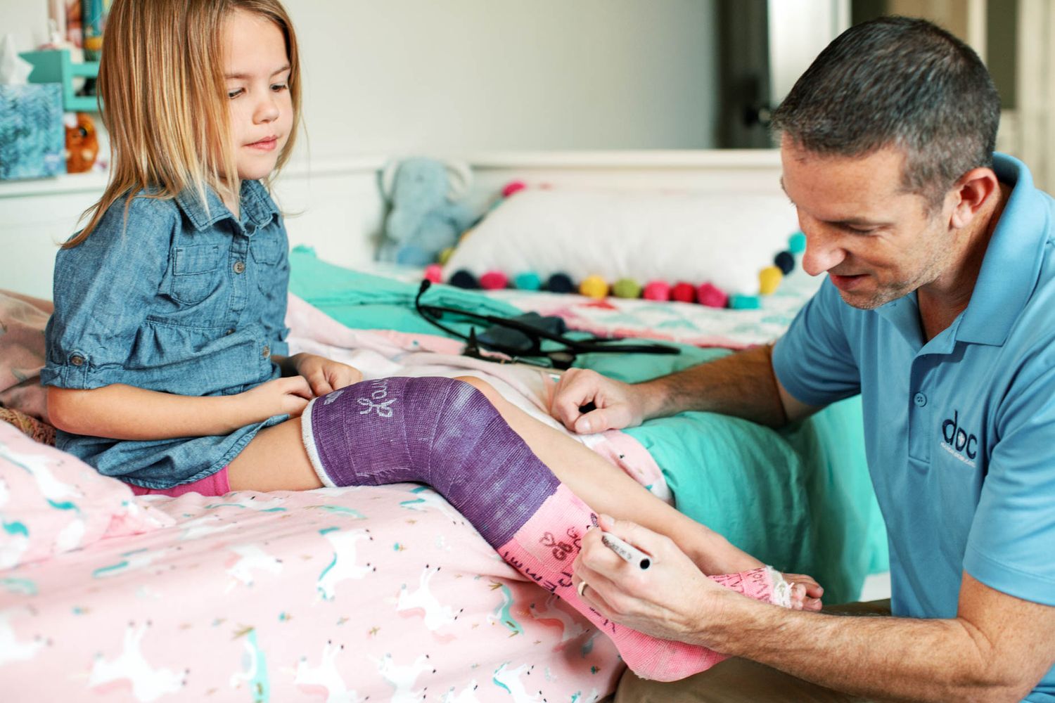 Man applying pink cast padding to a young girl's purple knee brace while she sits on a unicorn bedspread.