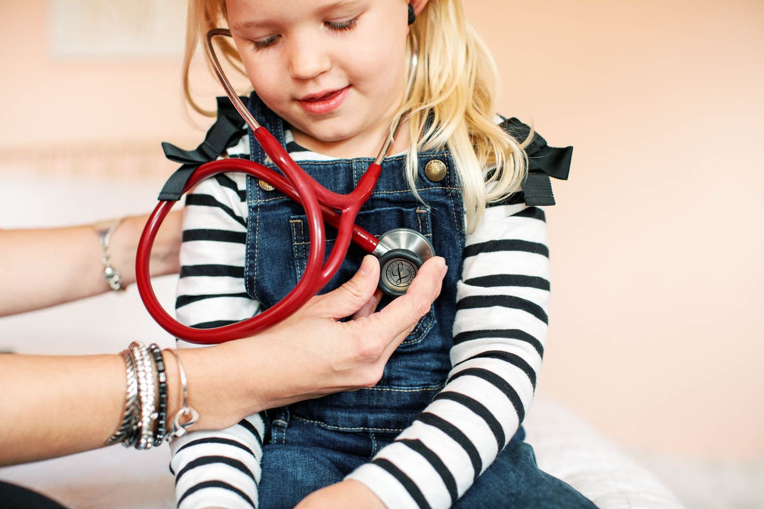 Doctor using red stethoscope to examine young blonde girl in striped shirt during pediatric checkup.