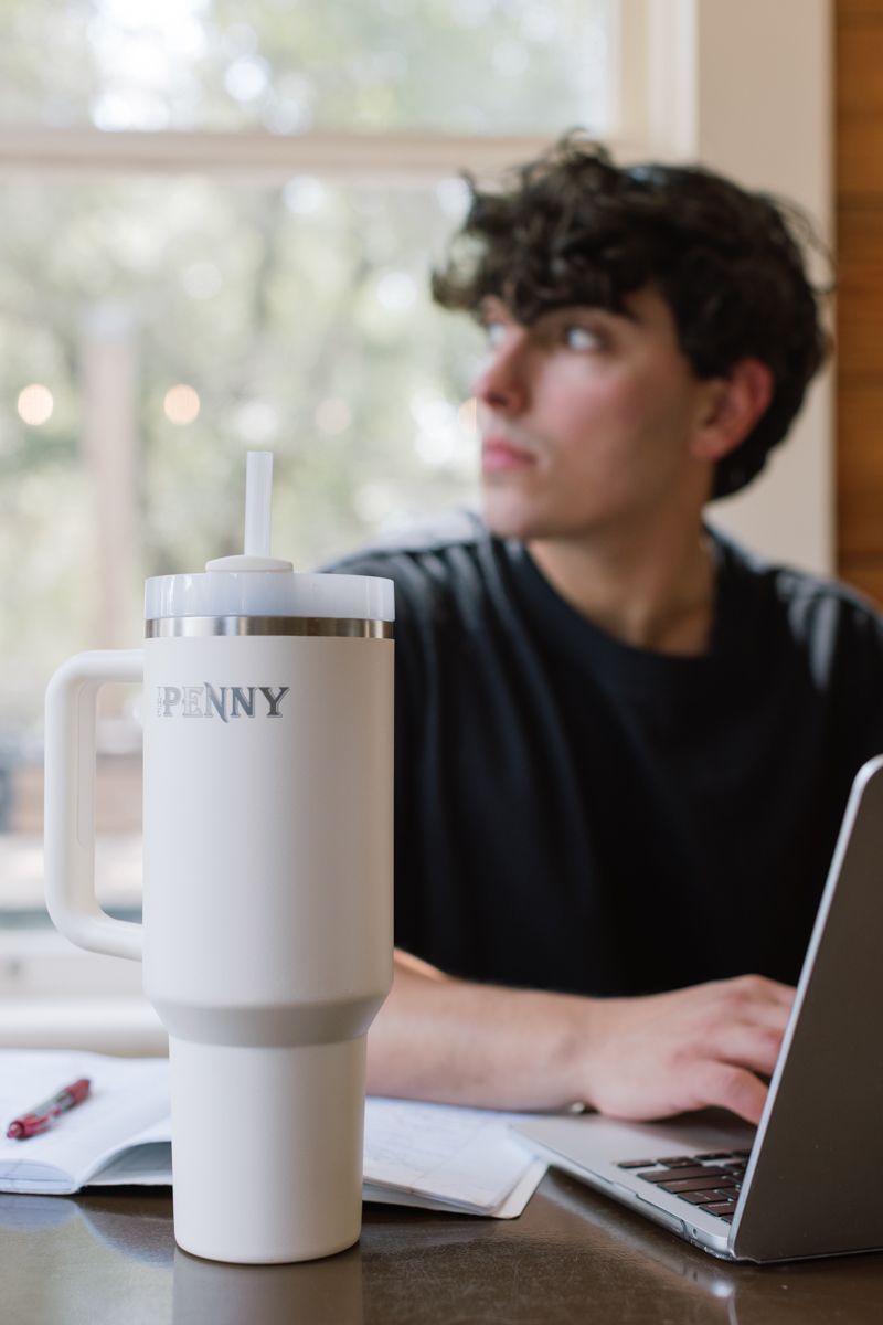 White Penny branded tumbler in foreground with young man working on laptop in background.