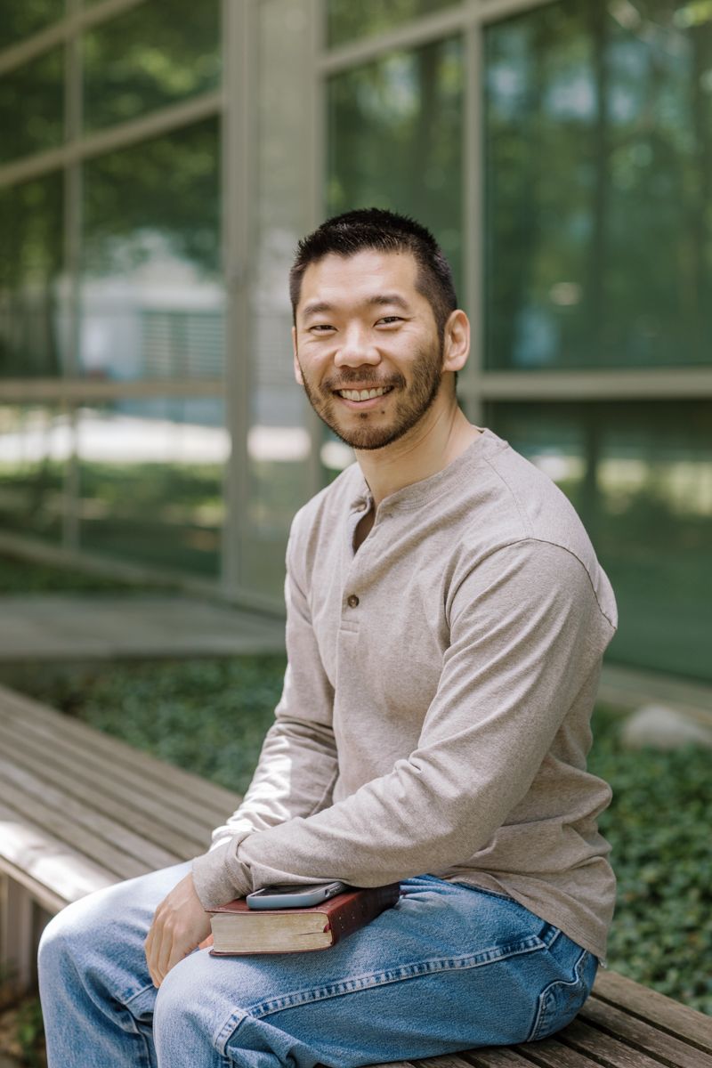 Smiling Asian man sitting on a bench outdoors holding a book, wearing a beige henley shirt.