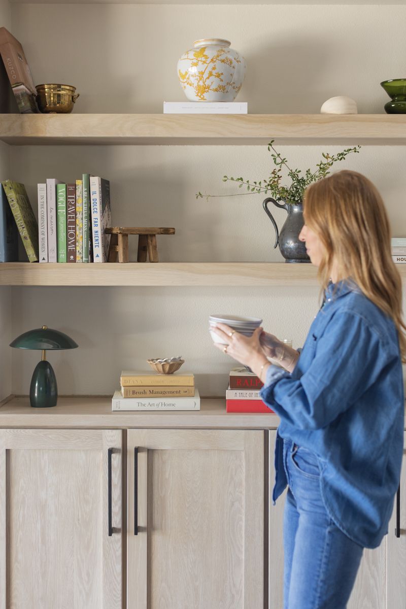 Woman in denim outfit arranging decor on natural wood floating shelves above light wood cabinetry.