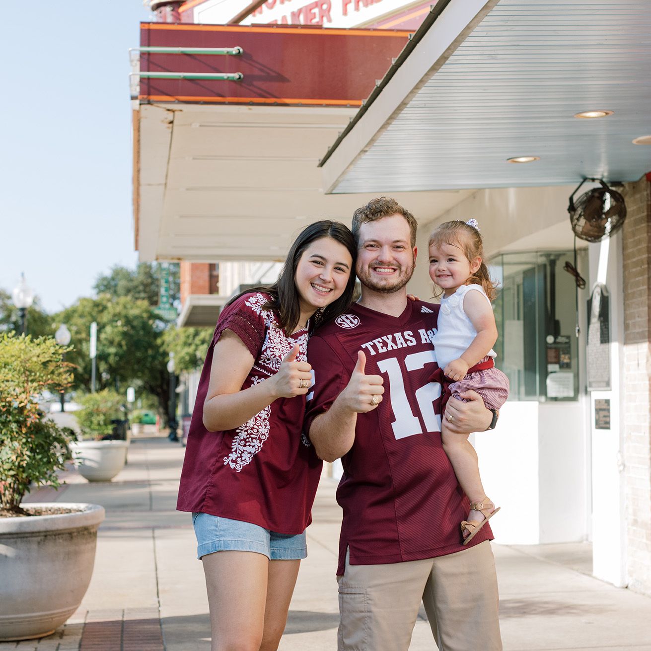 A young couple in Texas A&M jerseys smile and give thumbs up while holding a toddler on a sunny sidewalk.