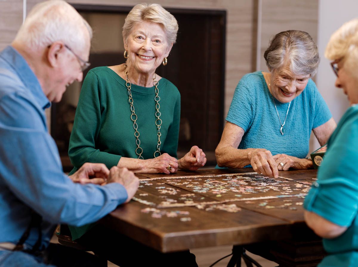 Four elderly adults smiling and working together on a jigsaw puzzle at a wooden table indoors.
