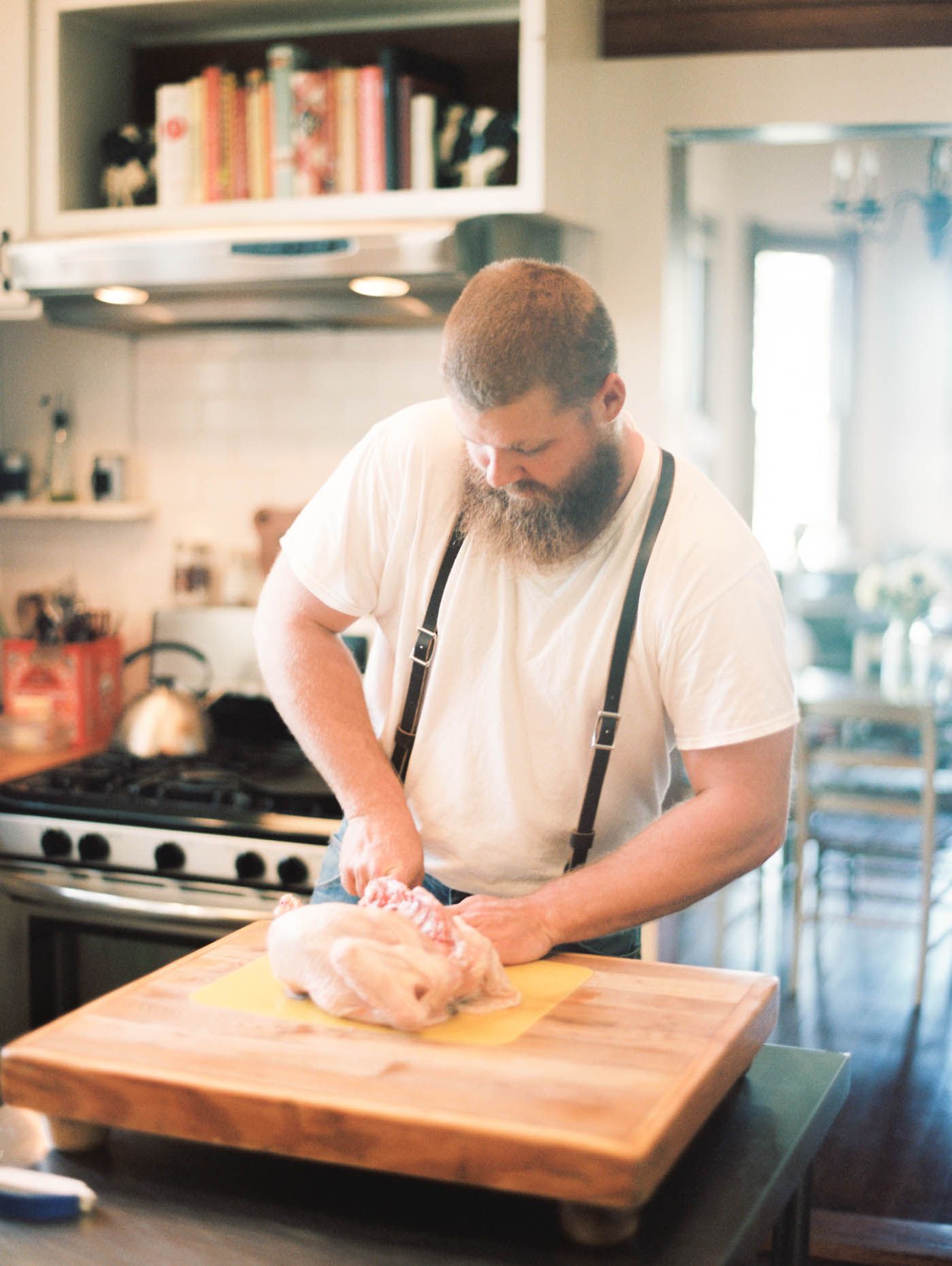 Bearded man in suspenders preparing raw chicken on wooden cutting board in kitchen.