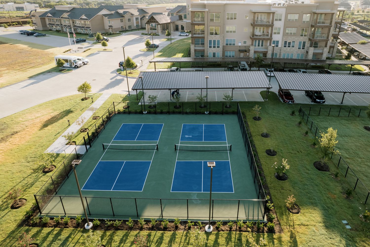 Aerial view of two blue pickleball courts surrounded by green grass and fencing near a modern residential apartment complex.
