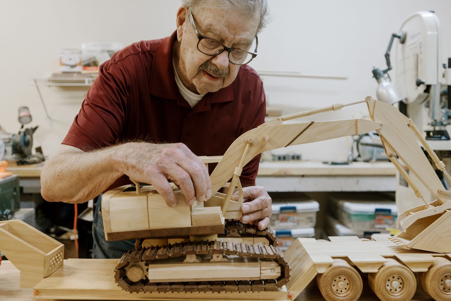 Elderly craftsman carefully assembling detailed wooden toy construction vehicles in a woodworking workshop.