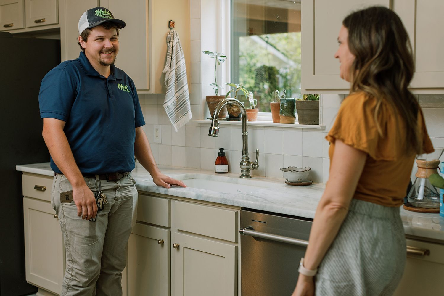 A plumber in a blue uniform discusses kitchen sink repairs with a homeowner in a bright, modern kitchen.