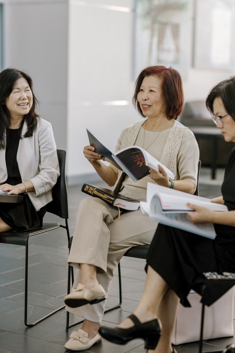 Three Asian women sitting in chairs, smiling and reading books together in a bright modern space.
